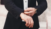 A person is adjusting the cuff of a tailored black suit jacket, wearing a white shirt underneath. The hands, adorned with a watch and a ring, are meticulously focused on the task, exuding a sense of elegance and sophistication.