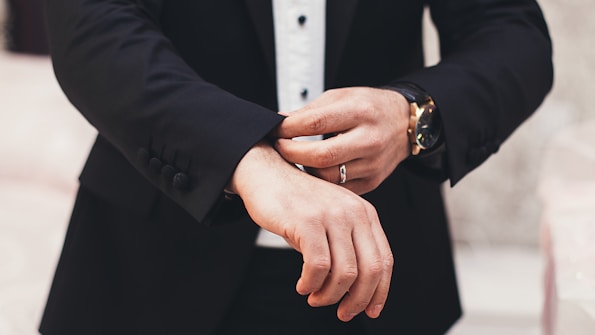 A person is adjusting the cuff of a tailored black suit jacket, wearing a white shirt underneath. The hands, adorned with a watch and a ring, are meticulously focused on the task, exuding a sense of elegance and sophistication.