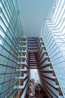 An architectural interior scene showcasing a multi-level atrium with geometric structures and expansive glass windows. The design is modern, featuring horizontal lines and a symmetrical viewpoint. People are visible on the ground floor, illustrating the scale and grandeur of the space.