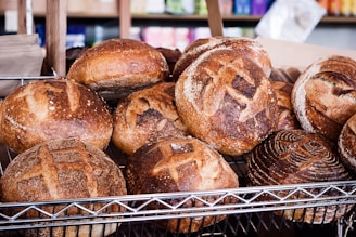 A display of freshly baked round loaves of bread with crusty, golden-brown exteriors arranged on a metal rack. The loaves have intricate scoring patterns and are set against a backdrop of assorted colorful packages.