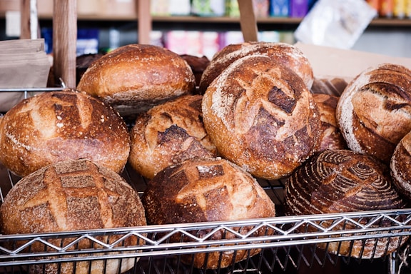 A display of freshly baked round loaves of bread with crusty, golden-brown exteriors arranged on a metal rack. The loaves have intricate scoring patterns and are set against a backdrop of assorted colorful packages.