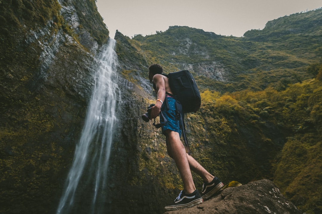man wearing blue shorts standing on rock formation while holding DSLR camera in front of waterfalls, Waterfall love
