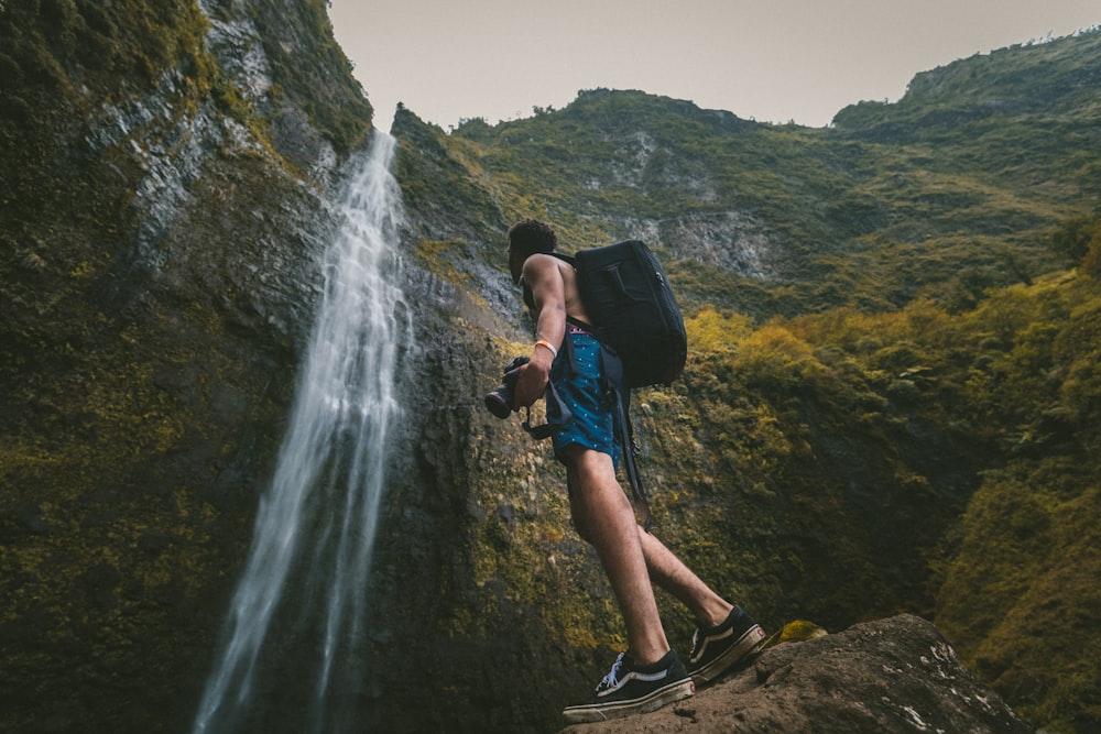 man wearing blue shorts standing on rock formation while holding DSLR camera in front of waterfalls