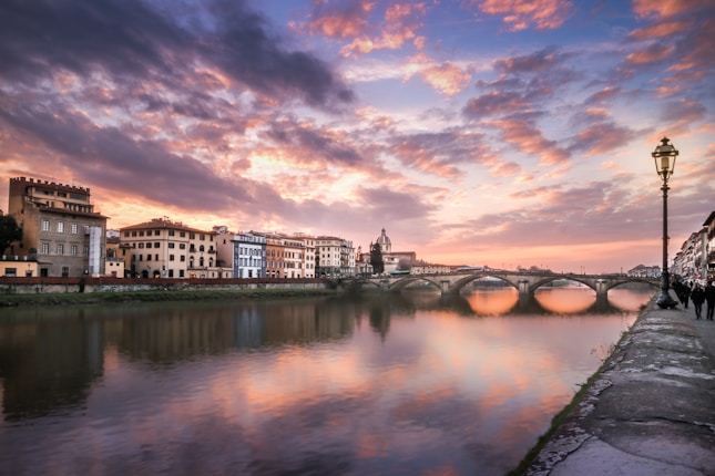 A picturesque cityscape of Florence at sunset, with colorful buildings lining the Arno River. A historic bridge stretches across the water, reflecting the captivating hues of the sky. Elegant streetlamps and people walking create a serene atmosphere.