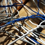 A close-up view of a tangle of twisted wires in blue, white, and gray, lying on a wooden surface, casting intricate shadows.