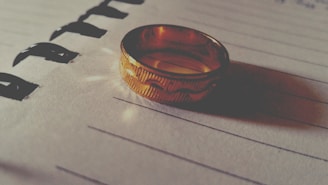 A gold ring lies on a lined paper, casting a shadow. The ring's texture and intricate design catch the light, creating a warm glow. Bold black lettering is partially visible, providing contrast with the soft surface of the paper.