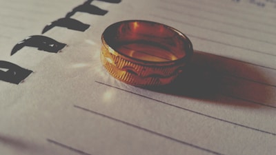 A gold ring lies on a lined paper, casting a shadow. The ring's texture and intricate design catch the light, creating a warm glow. Bold black lettering is partially visible, providing contrast with the soft surface of the paper.