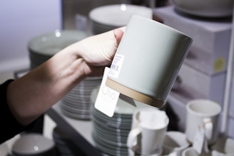 A hand holding a ceramic mug with a light tan base and a light gray body. The mug has a label attached, and there are several stacks of similar mugs and dishes in the background on store shelves.