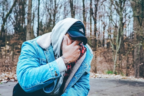 A person dressed in a denim jacket with a fleece lining and a hoodies stands outdoors. They are wearing a black cap and have their hand covering part of their face. The background is a forest with bare trees and fallen leaves on the ground, suggesting a chilly autumn or winter setting.