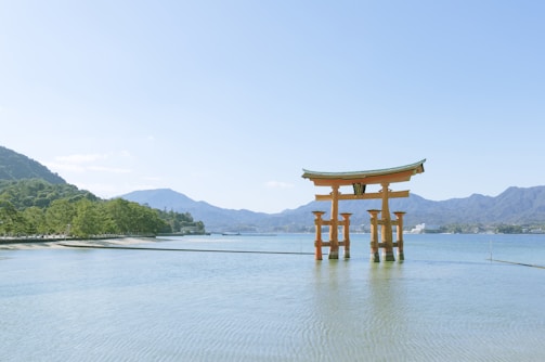 A traditional Japanese torii gate stands in shallow water against a backdrop of blue sky and distant mountains. Surrounding the area are green trees lining the shore, with gentle ripples visible in the calm water.