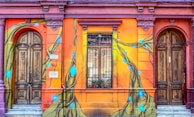 A vibrant street mural covers the exterior of an ornate building with two wooden doors. The walls are painted with bright orange and red hues, showcasing a dynamic design featuring flowing lines and blue glowing orbs. The artwork intricately weaves around the architectural elements, including a window with bars and decorative moldings.