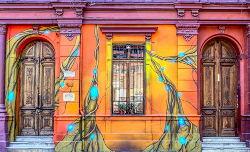 A vibrant street mural covers the exterior of an ornate building with two wooden doors. The walls are painted with bright orange and red hues, showcasing a dynamic design featuring flowing lines and blue glowing orbs. The artwork intricately weaves around the architectural elements, including a window with bars and decorative moldings.