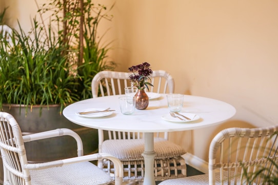A round white table is set with two plates and minimal cutlery, surrounded by white wicker chairs. In the center of the table is a small vase with purple flowers. The background features a large planter filled with tall green plants against a light-colored wall, creating a serene and cozy atmosphere.