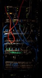A dimly lit server rack filled with network equipment. Multiple colored cables, predominantly red, blue, and black, are neatly organized and connected to various devices such as switches and routers. Small LED lights on the equipment indicate active connections.