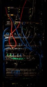 A dimly lit server rack filled with network equipment. Multiple colored cables, predominantly red, blue, and black, are neatly organized and connected to various devices such as switches and routers. Small LED lights on the equipment indicate active connections.