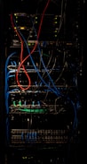 A dimly lit server rack filled with network equipment. Multiple colored cables, predominantly red, blue, and black, are neatly organized and connected to various devices such as switches and routers. Small LED lights on the equipment indicate active connections.