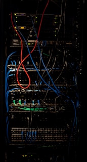 A dimly lit server rack filled with network equipment. Multiple colored cables, predominantly red, blue, and black, are neatly organized and connected to various devices such as switches and routers. Small LED lights on the equipment indicate active connections.