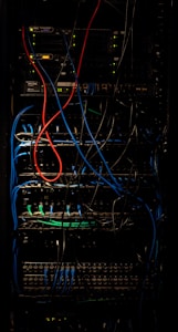 A dimly lit server rack filled with network equipment. Multiple colored cables, predominantly red, blue, and black, are neatly organized and connected to various devices such as switches and routers. Small LED lights on the equipment indicate active connections.
