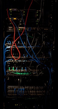 A dimly lit server rack filled with network equipment. Multiple colored cables, predominantly red, blue, and black, are neatly organized and connected to various devices such as switches and routers. Small LED lights on the equipment indicate active connections.