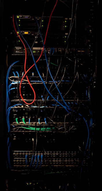 A dimly lit server rack filled with network equipment. Multiple colored cables, predominantly red, blue, and black, are neatly organized and connected to various devices such as switches and routers. Small LED lights on the equipment indicate active connections.