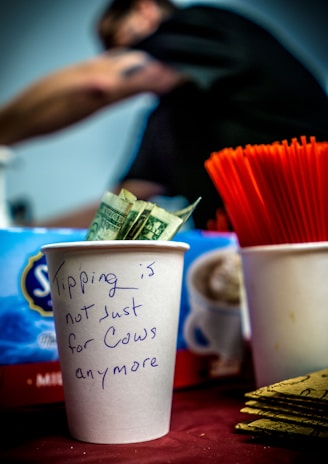 A white paper cup filled with dollar bills is prominently displayed. The cup has a handwritten message that reads, 'Tipping is not just for cows anymore.' In the background, there’s a red container with straws and a blurred figure of a person.