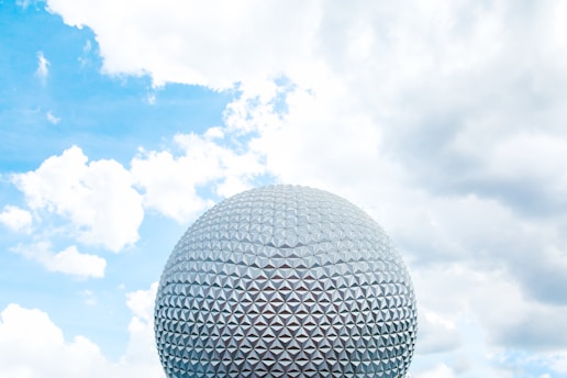 A large geodesic sphere structure is set against a backdrop of a partly cloudy sky with bright blue hues. The sphere is composed of triangular panels, giving it a futuristic appearance.