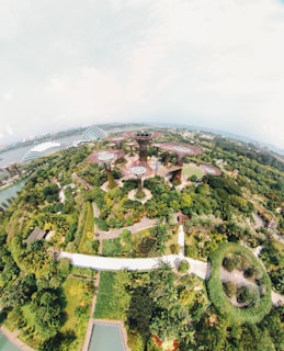 An aerial view of a lush green park featuring multiple tall, tree-like structures with canopies. The landscape includes pathways surrounded by diverse vegetation and water bodies. Futuristic architectural elements can be seen in the background.