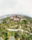 An aerial view of a lush green park featuring multiple tall, tree-like structures with canopies. The landscape includes pathways surrounded by diverse vegetation and water bodies. Futuristic architectural elements can be seen in the background.