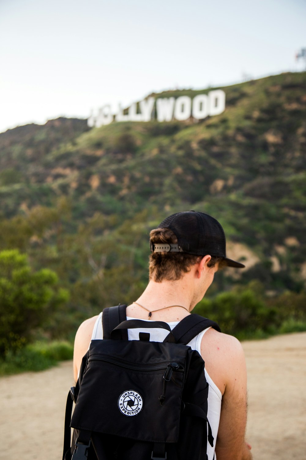man wearing white tank top and black snapback cap carrying black backpack in distance hollywood signboard at daytime