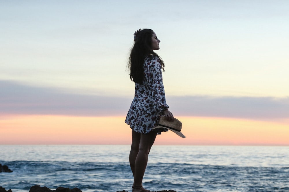 Mujer sosteniendo sombrero marrón para el sol en la orilla durante la puesta del sol