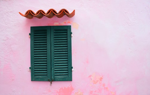 A closed, green wooden shutter is set against a light pink, textured wall. Above the shutter is a small, decorative wavy metal awning, painted in orange.