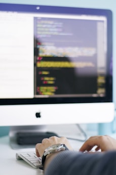 A person is typing on a keyboard in front of a computer screen displaying code or a programming interface. The environment suggests a focused and professional atmosphere.