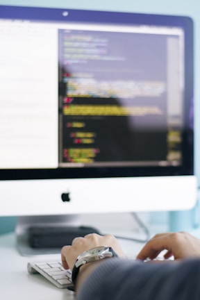 A person is typing on a keyboard in front of a computer screen displaying code or a programming interface. The environment suggests a focused and professional atmosphere.