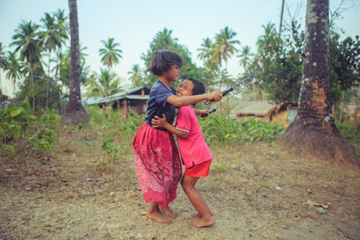 Two children are playing outdoors in a rural area surrounded by palm trees and greenery. They appear to be happy and engaged in playful interaction, possibly pretending to fish with a makeshift rod.