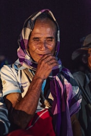 An elderly person is sitting in a dark environment, wearing a patterned headscarf with purple and white colors. The individual has a gentle smile and expressive eyes, exuding a sense of wisdom and calmness. The attire consists of traditional clothing with text embroidered on the shirt.