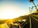 A convertible car travels down a palm-lined coastal road during sunset. Passengers in the vehicle appear to be enjoying the ride. The sky is brightly lit by the setting sun, casting a warm glow over the scene.