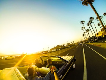 A convertible car travels down a palm-lined coastal road during sunset. Passengers in the vehicle appear to be enjoying the ride. The sky is brightly lit by the setting sun, casting a warm glow over the scene.