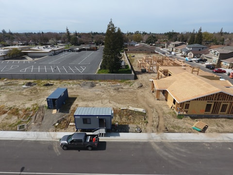 Aerial view of a construction site with a partially built house on the right side and a blue shipping container on the left. There is also a truck parked on the street. In the background, there is a parking lot with empty spaces, trees, and several residential houses.