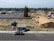 Aerial view of a construction site with a partially built house on the right side and a blue shipping container on the left. There is also a truck parked on the street. In the background, there is a parking lot with empty spaces, trees, and several residential houses.