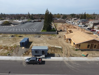 Aerial view of a construction site with a partially built house on the right side and a blue shipping container on the left. There is also a truck parked on the street. In the background, there is a parking lot with empty spaces, trees, and several residential houses.