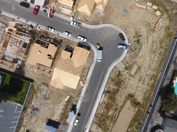An aerial view of a suburban area under development with several houses under construction, surrounded by a mixture of paved roads and dirt areas. Visible are wooden frameworks of partially built houses, completed roofs, and various construction materials scattered around. Several vehicles are parked along the roads, including trucks and cars.