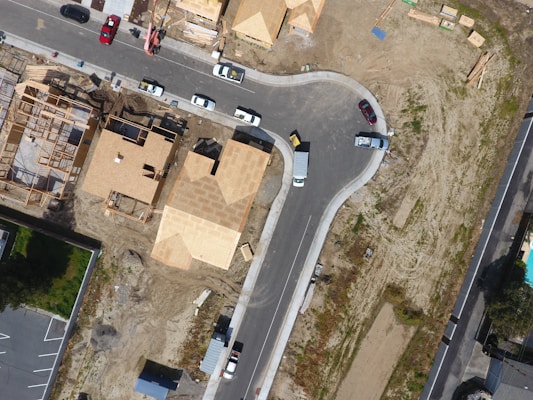 An aerial view of a suburban area under development with several houses under construction, surrounded by a mixture of paved roads and dirt areas. Visible are wooden frameworks of partially built houses, completed roofs, and various construction materials scattered around. Several vehicles are parked along the roads, including trucks and cars.