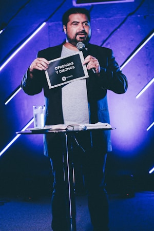 A person stands on stage holding a sign that reads 'Ofrendas y Diezmos.' They are speaking into a microphone, and there is a small table in front with a glass of water and some papers. The background features blue and purple lighting with diagonal light bars.