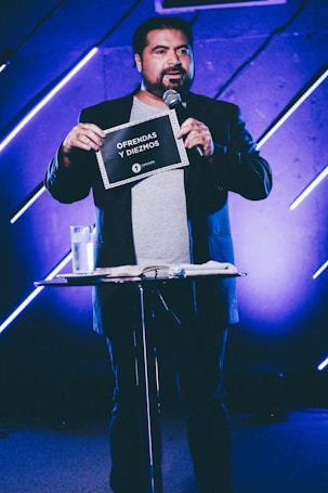 A person stands on stage holding a sign that reads 'Ofrendas y Diezmos.' They are speaking into a microphone, and there is a small table in front with a glass of water and some papers. The background features blue and purple lighting with diagonal light bars.