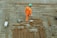 A construction worker in an orange uniform and a green helmet is seen working on a construction site. The ground is covered with a grid of steel rebar and various tools, including a power drill and cables, are scattered around.