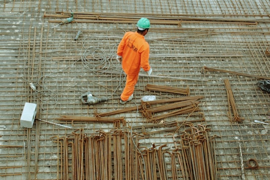 A construction worker in an orange uniform and a green helmet is seen working on a construction site. The ground is covered with a grid of steel rebar and various tools, including a power drill and cables, are scattered around.
