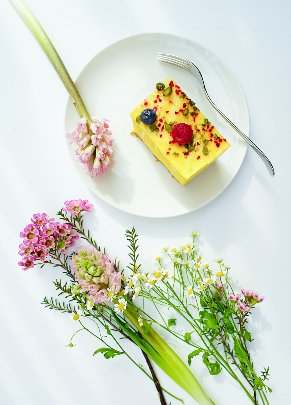 An overhead shot of a piece of cheesecake on a plate next to small bouquets of various flowers