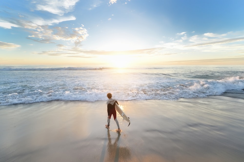 uomo che tiene la tavola da surf che cammina sulla riva durante il giorno