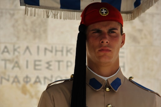 A guard dressed in traditional military uniform stands solemnly. The uniform includes a red beret, beige overcoat with blue and gold accents. The background features a stone wall with inscriptions.