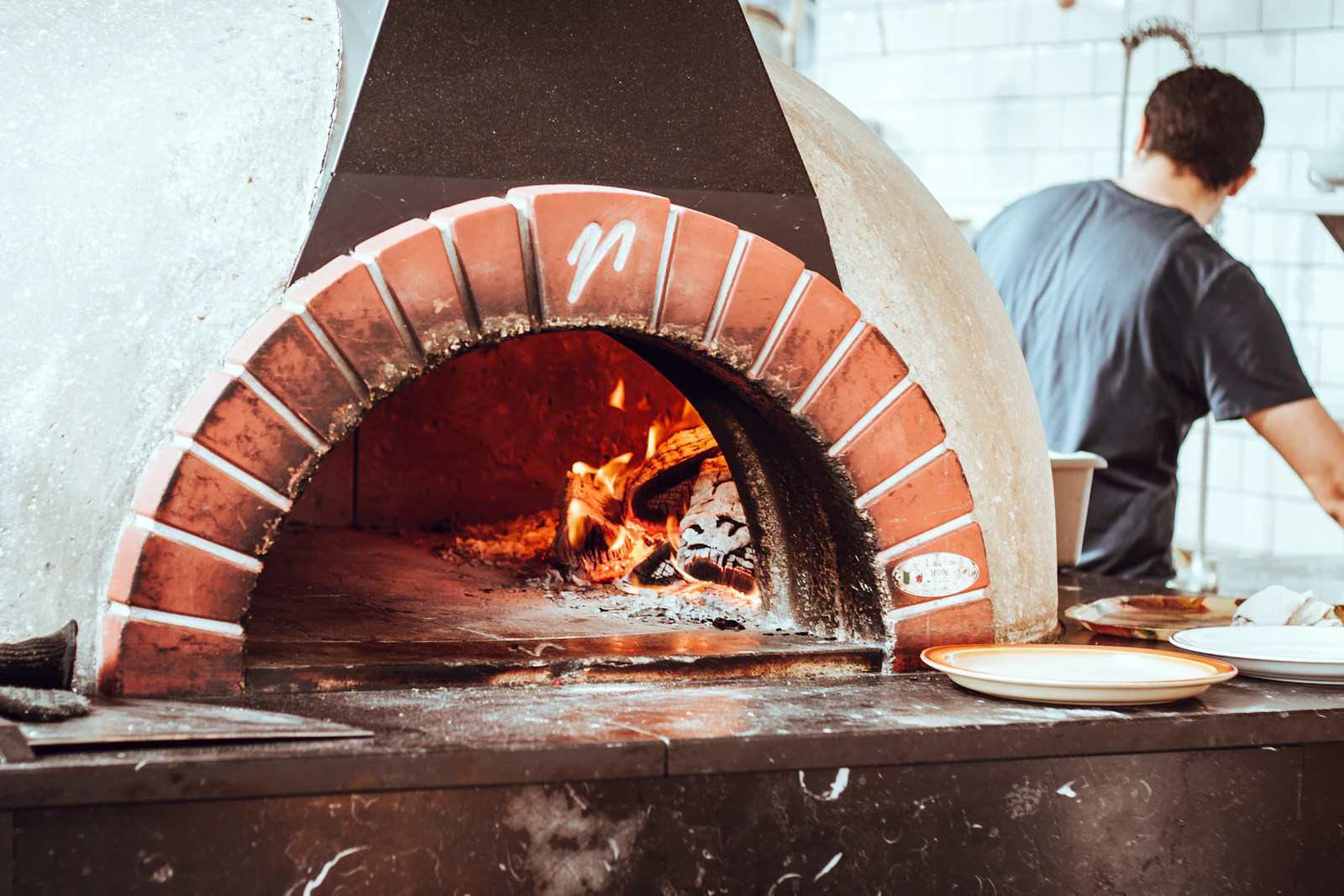 The open mouth of a brick oven with the fire lit inside, a cook working alongside it
